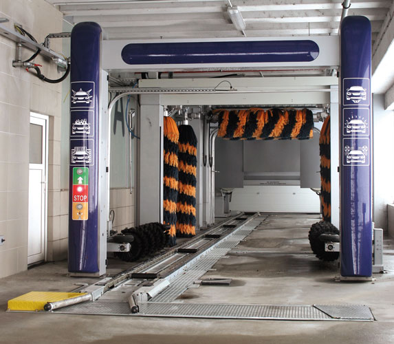 A car wash station featuring large rotating brushes, with a stop button and signage on the sides. The area is clean and well-lit.