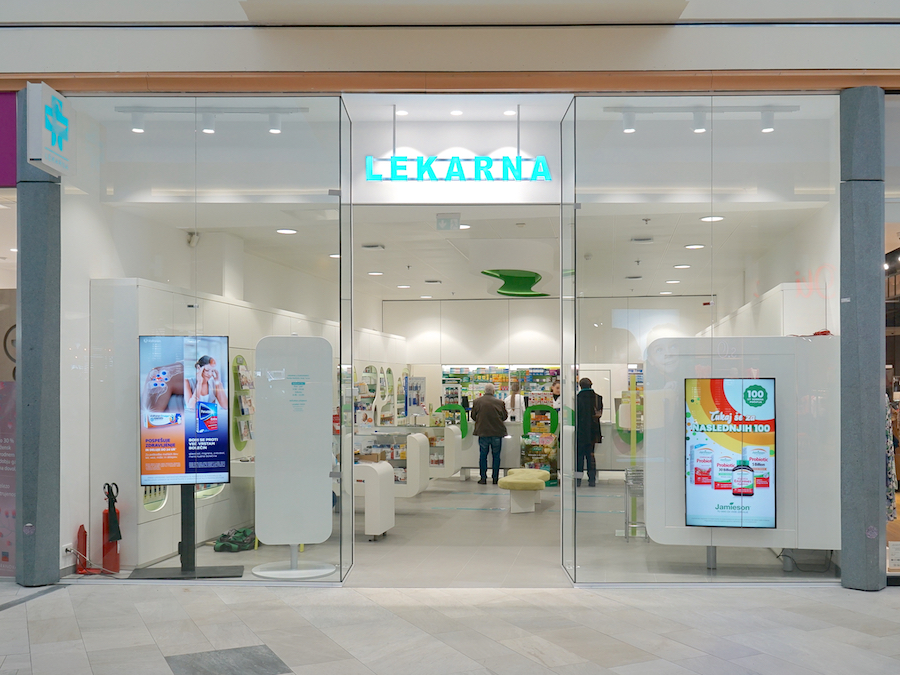 A modern pharmacy entrance with glass doors, featuring bright signage and displays of health products inside.