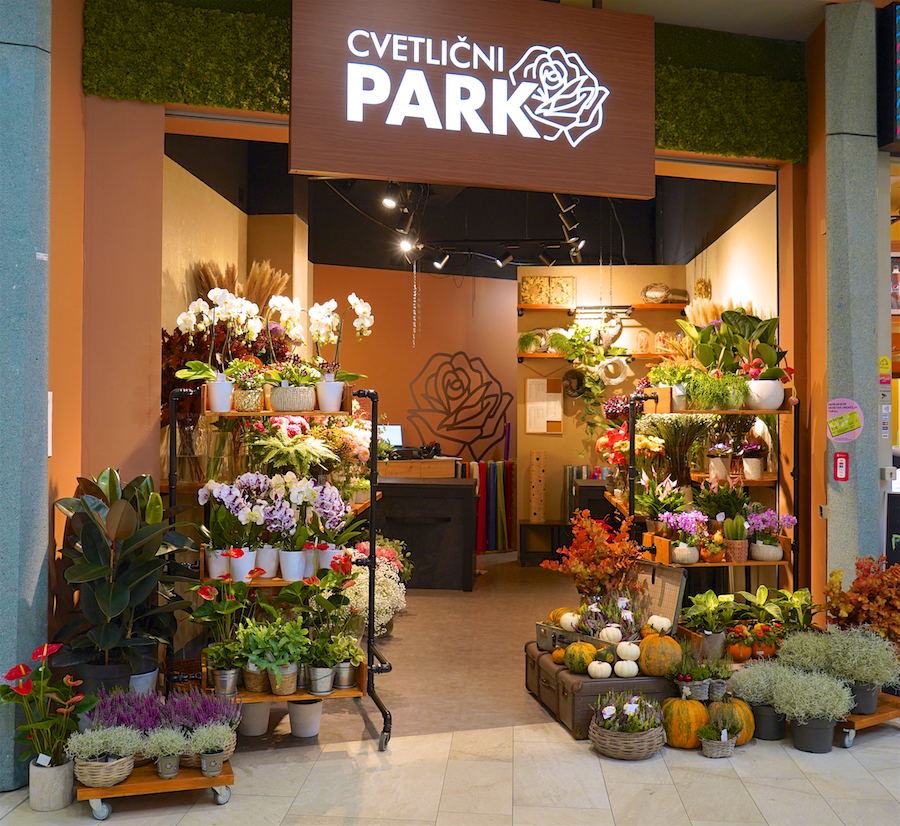 A flower shop entrance with various plants and flowers displayed, featuring a sign that reads "Cvetlični Park."