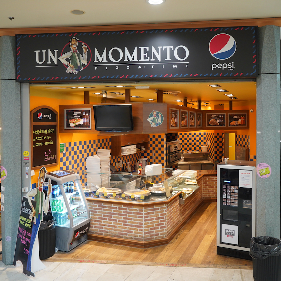 A pizza shop named "Un Momento" featuring a colorful interior, display cases, and a Pepsi logo.