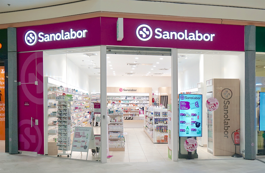 A brightly lit pharmacy storefront with the name "Sanolabor" prominently displayed. Shelves filled with products are visible inside.