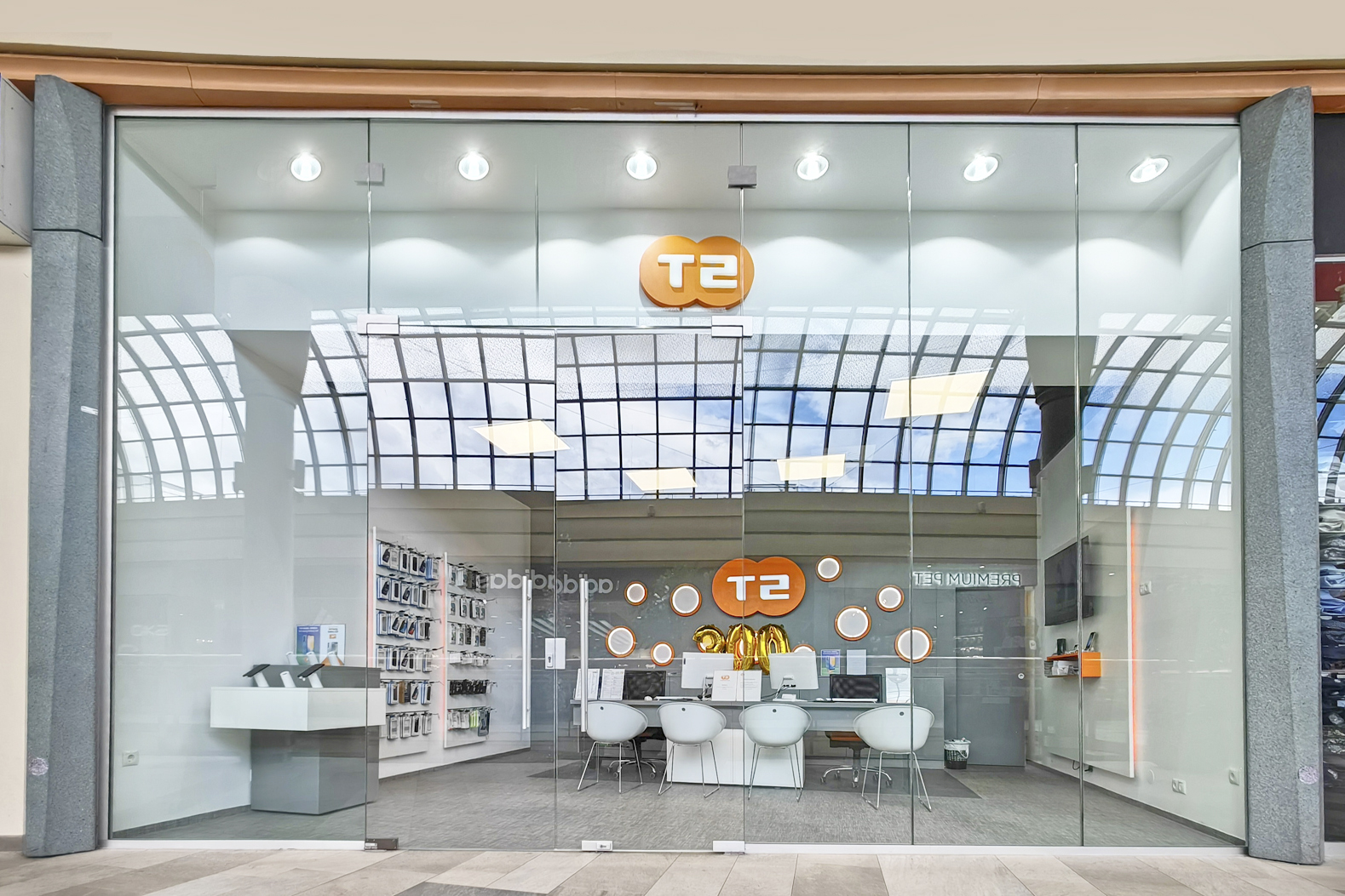 A modern retail store with glass doors, featuring the "T2" logo, displays tech products and seating inside.