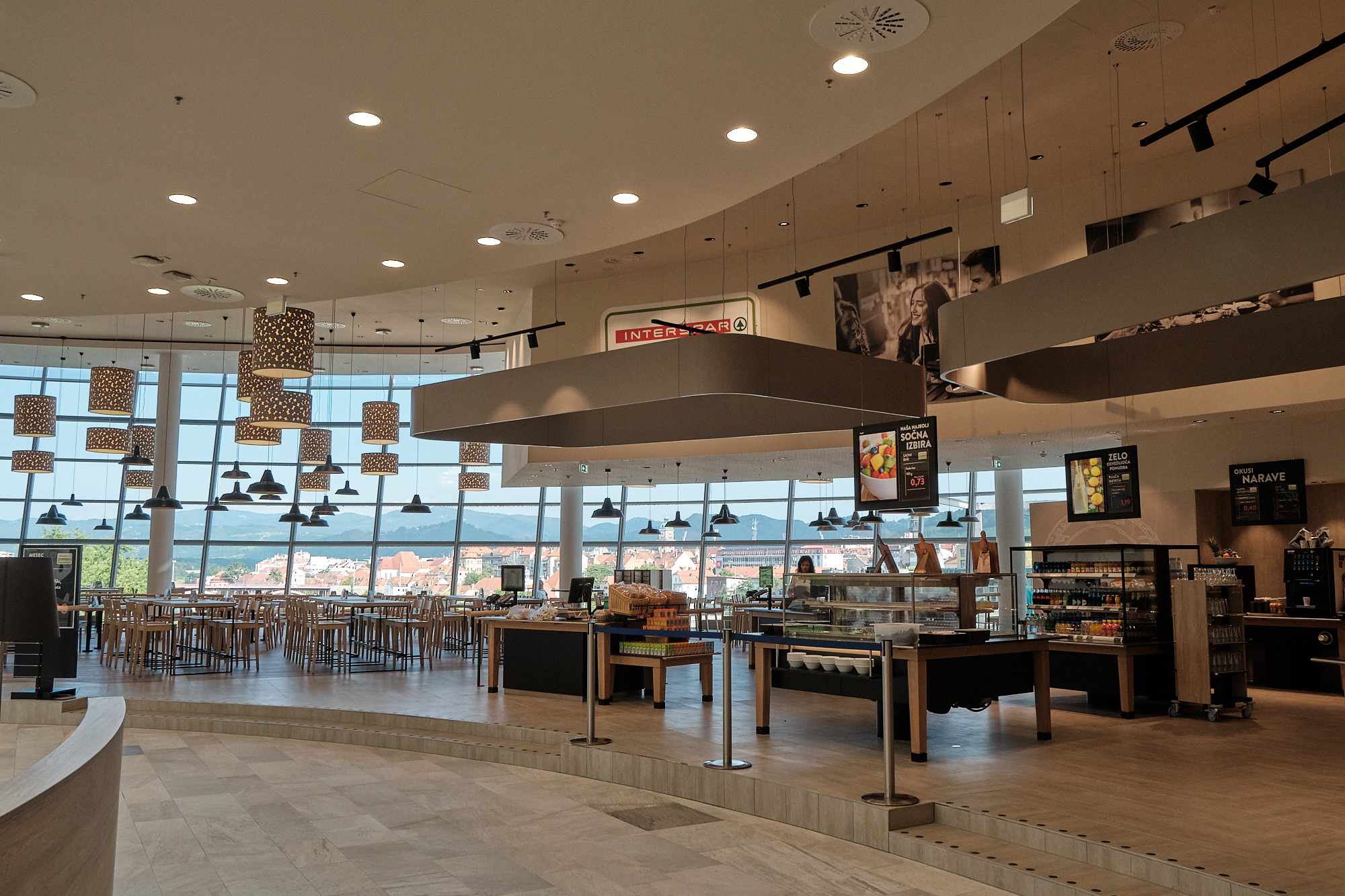 A modern café interior with large windows, wooden tables, and hanging lights. Displays of food and drinks are visible.