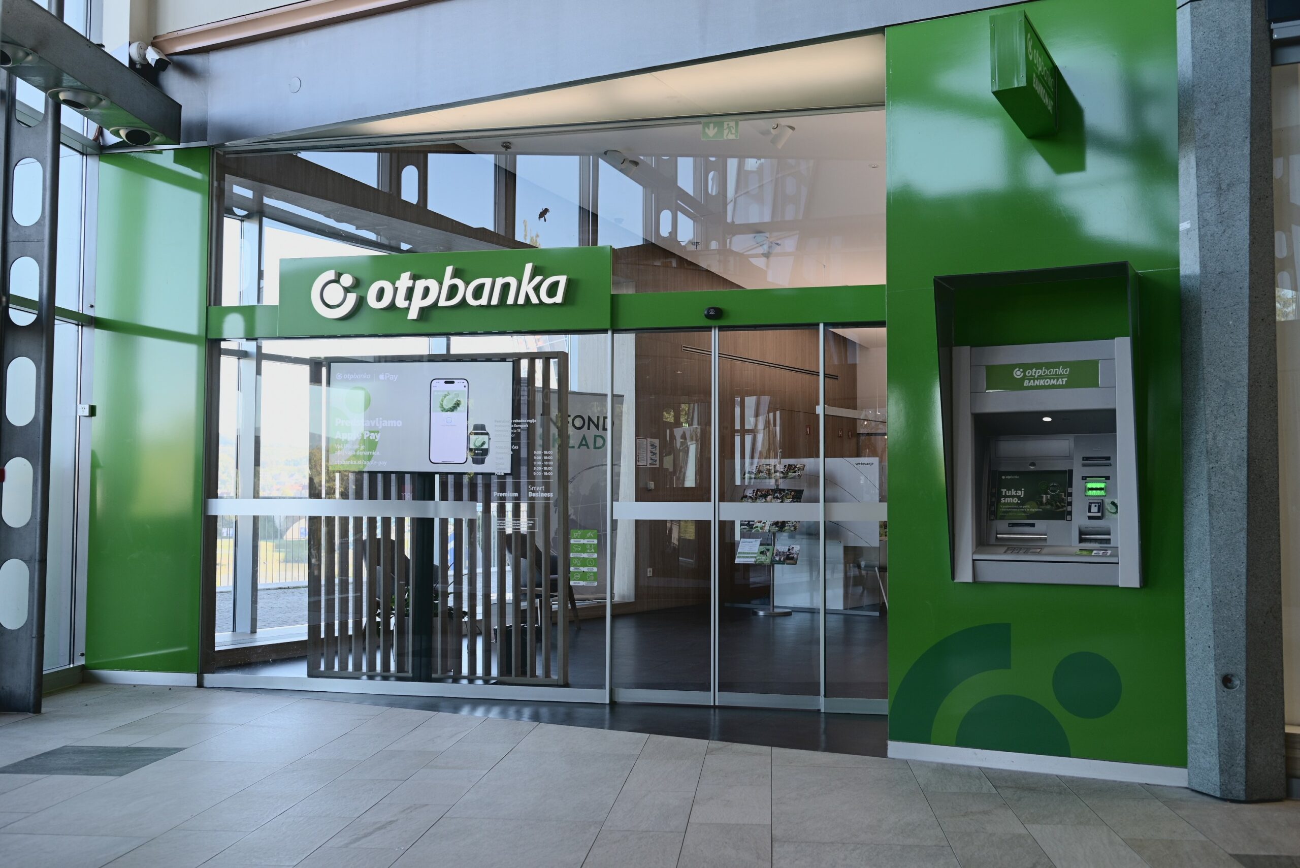 Entrance of an OTP Bank branch featuring a green logo and an ATM outside. Modern design with glass doors and promotional displays.