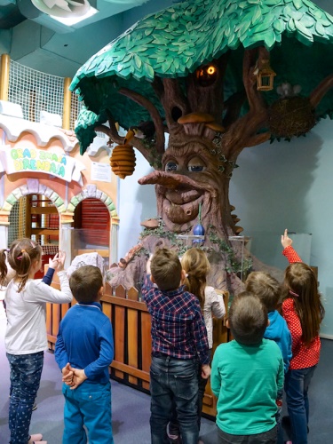 A group of children is pointing at a whimsical tree sculpture with a face, surrounded by playful decorations.