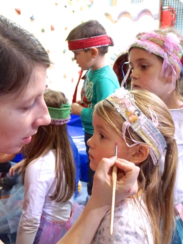 A child is getting face paint applied by an adult, while other children are in the background, wearing colorful headbands.