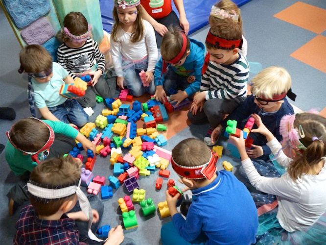 A group of children wearing blindfolds play with colorful building blocks on the floor, engaged in a collaborative activity.