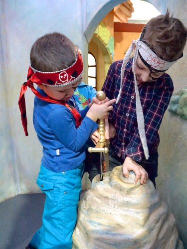 Two children dressed as pirates are playfully trying to pull a sword from a rock in a colorful indoor setting.