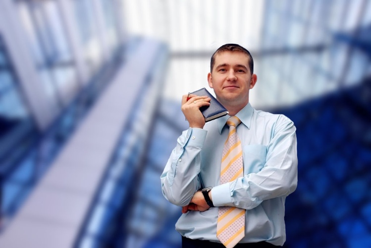 A businessman in a light blue shirt and striped tie stands confidently, holding a notebook, with a modern glass building in the background.