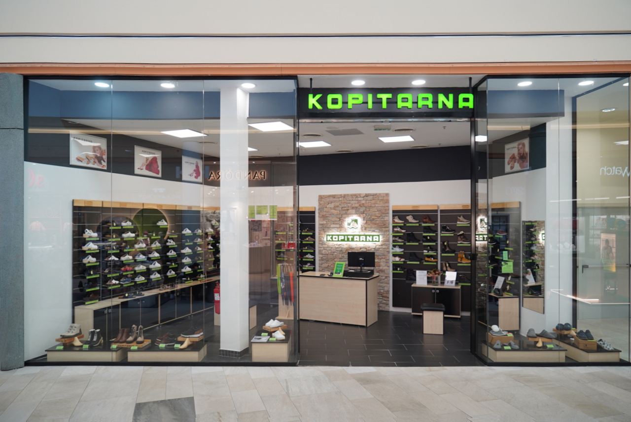 A shoe store named "Kopitarna" with a modern design, featuring various shoes displayed on shelves and a checkout area.