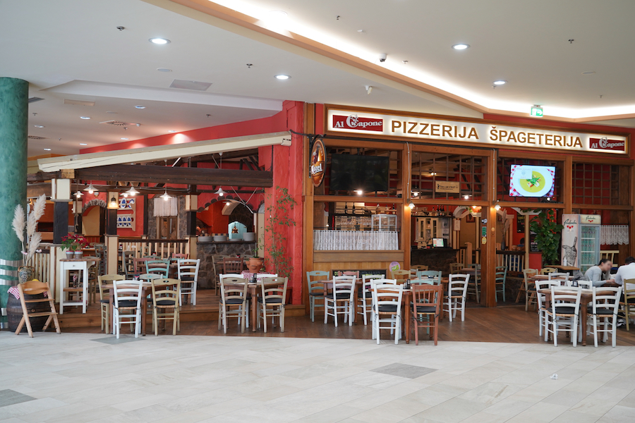 A cozy restaurant interior featuring wooden tables and chairs, with a sign reading "Pizzerija Špageterija" and a warm, inviting atmosphere.