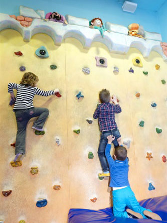 Three children are climbing a colorful indoor rock wall, with one child near the top and two others lower down.