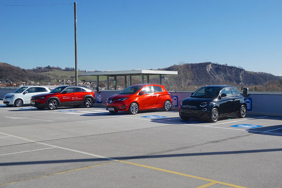 A parking lot with four cars: a white car, a red car, a black car, and a Mazda SUV, against a clear blue sky and hills in the background.