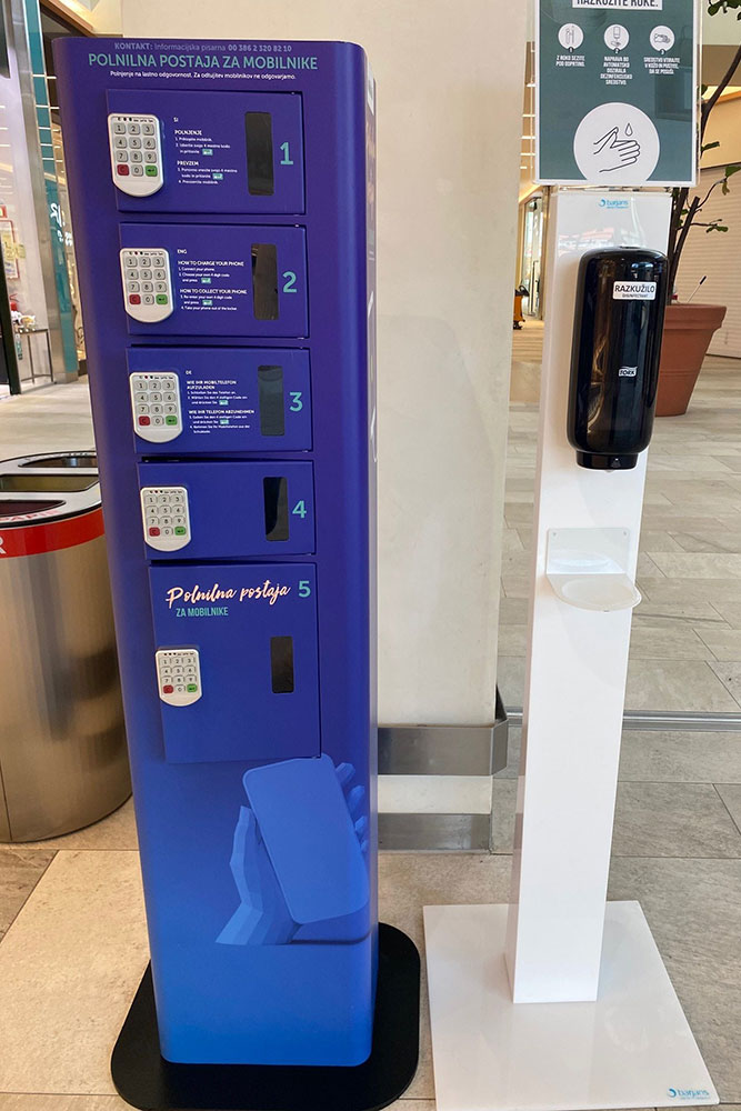 A purple kiosk with multiple compartments for mobile devices and a hand sanitizer dispenser nearby.