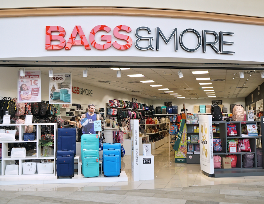 A retail store named "BAGS & MORE" featuring various bags and luggage, with promotional signs and a life-size cutout.