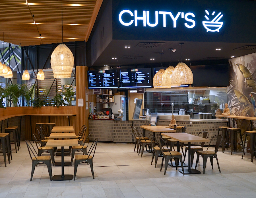 A modern restaurant interior featuring wooden decor, hanging lights, and a menu display. Tables and chairs are arranged for dining.