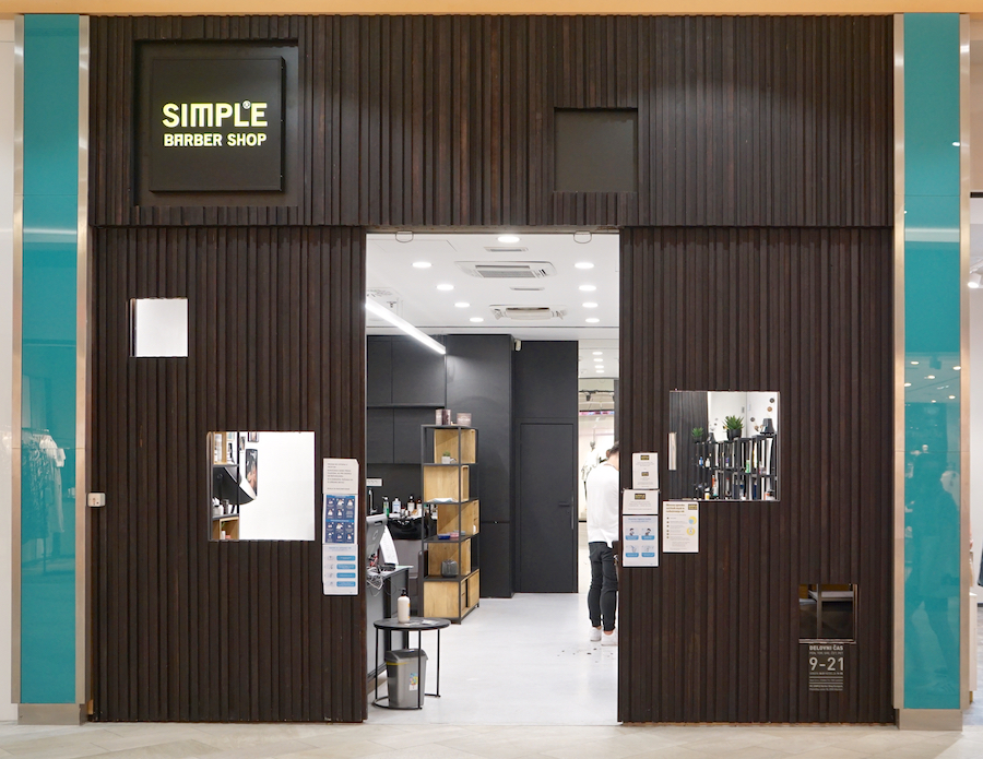 A modern barber shop entrance with a wooden facade, featuring a sign that reads "SIMPLE BARBER SHOP." Inside, there are shelves and a stylist at work.