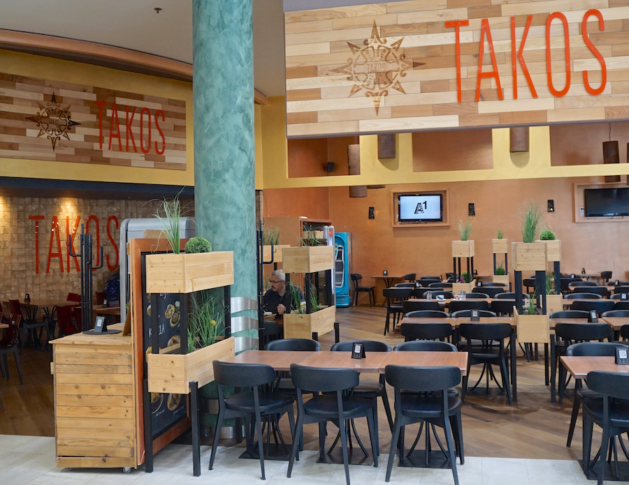 A modern restaurant interior featuring wooden decor, multiple tables, and the word "TAKOS" prominently displayed on the walls.