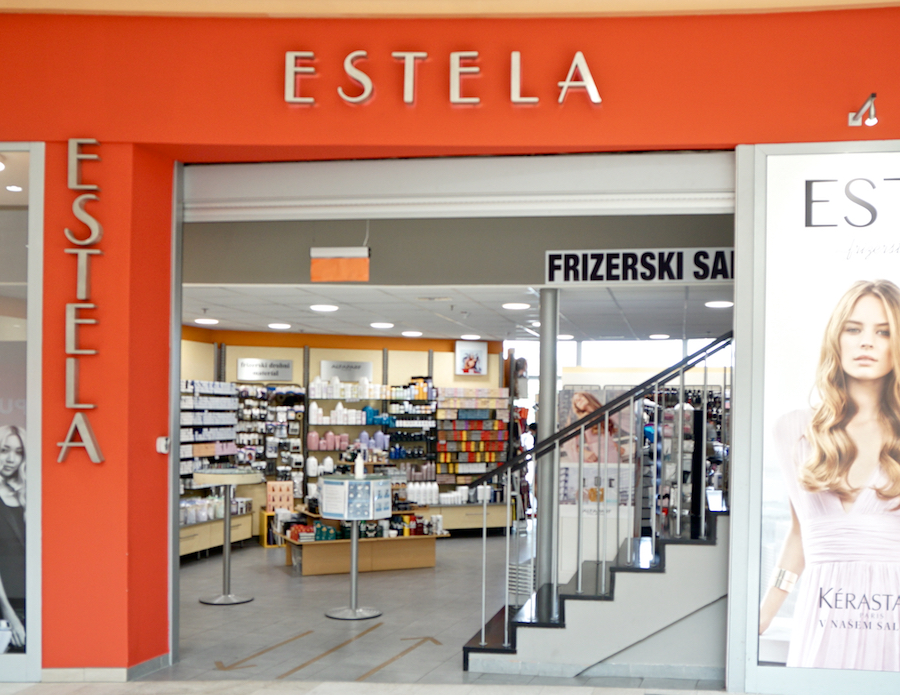 A storefront labeled "ESTELA" with a hair salon sign. Inside, shelves display various beauty products. Stairs lead to an upper level.