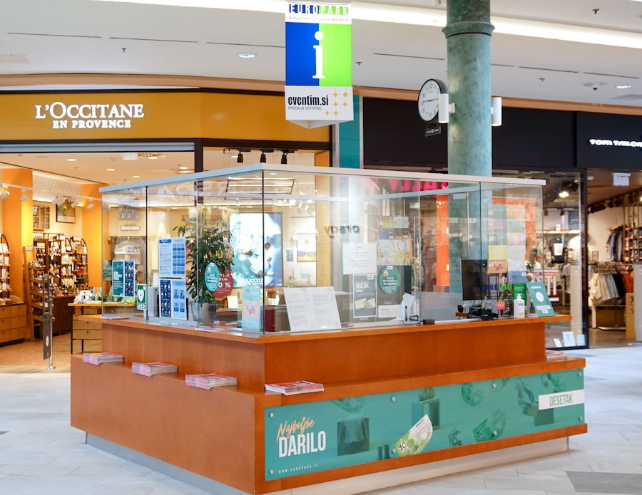 A customer service kiosk in a shopping mall, featuring a glass enclosure and signage for nearby stores, including L'Occitane.