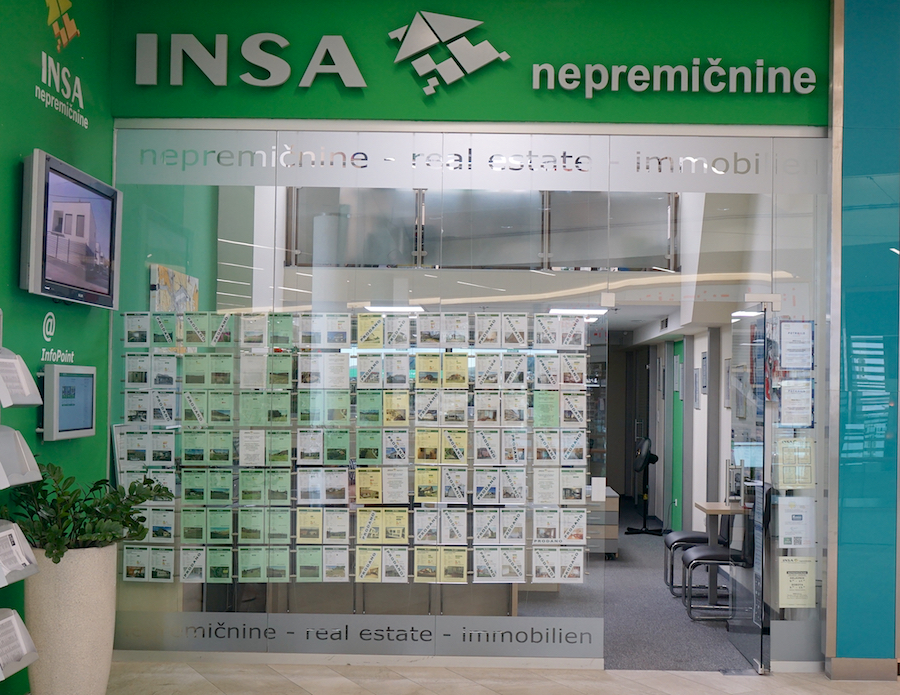 A real estate office with a green sign displaying "INSA" and listings on a glass wall. There are informational displays and a TV.