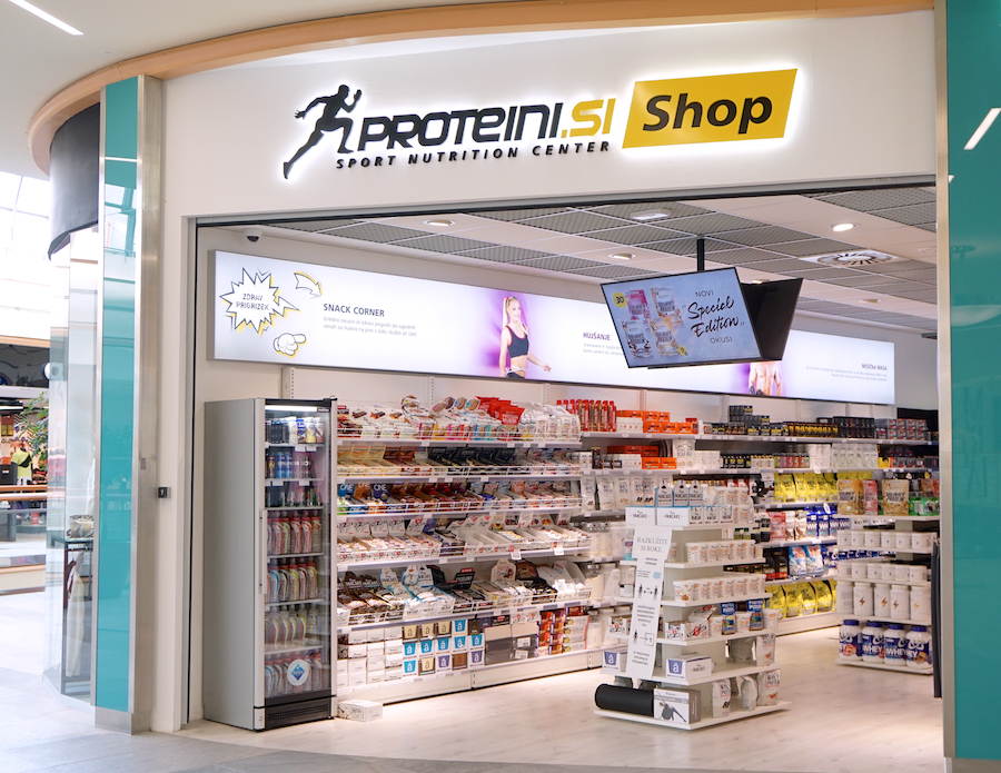 A storefront for "Proteini.si Shop," a sport nutrition center, featuring shelves stocked with various health products and a digital display.