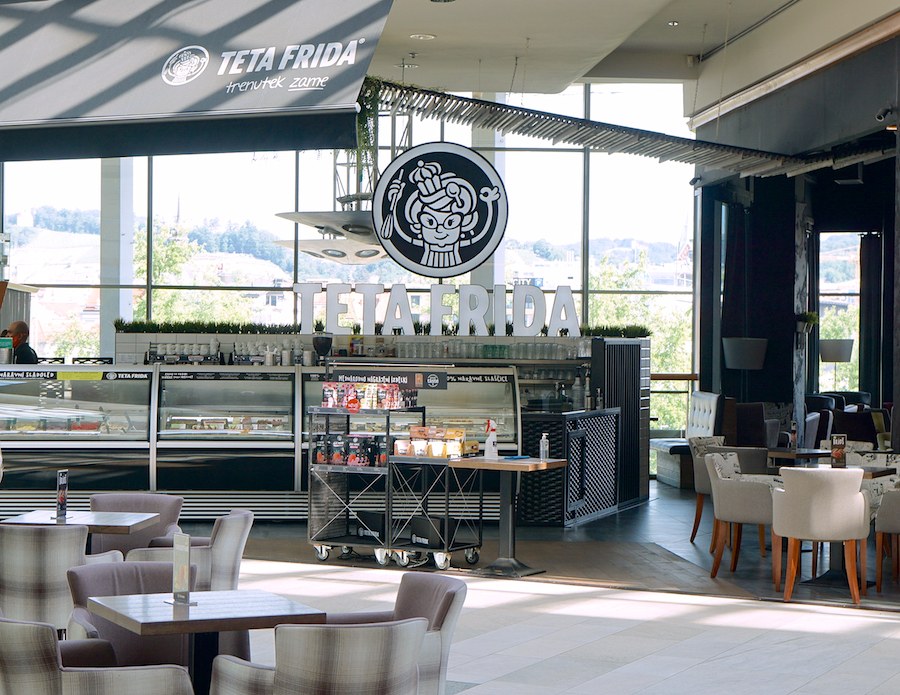 A modern café interior featuring a large sign for "TETA FRIDA," with seating areas and a display of desserts.