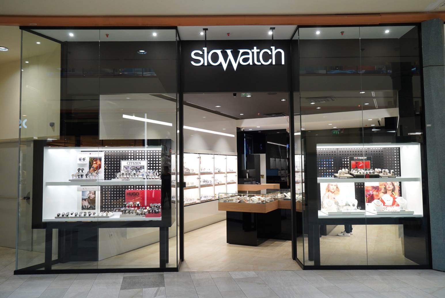 A modern watch store with glass displays showcasing various watches and accessories, featuring the "slowatch" sign prominently.