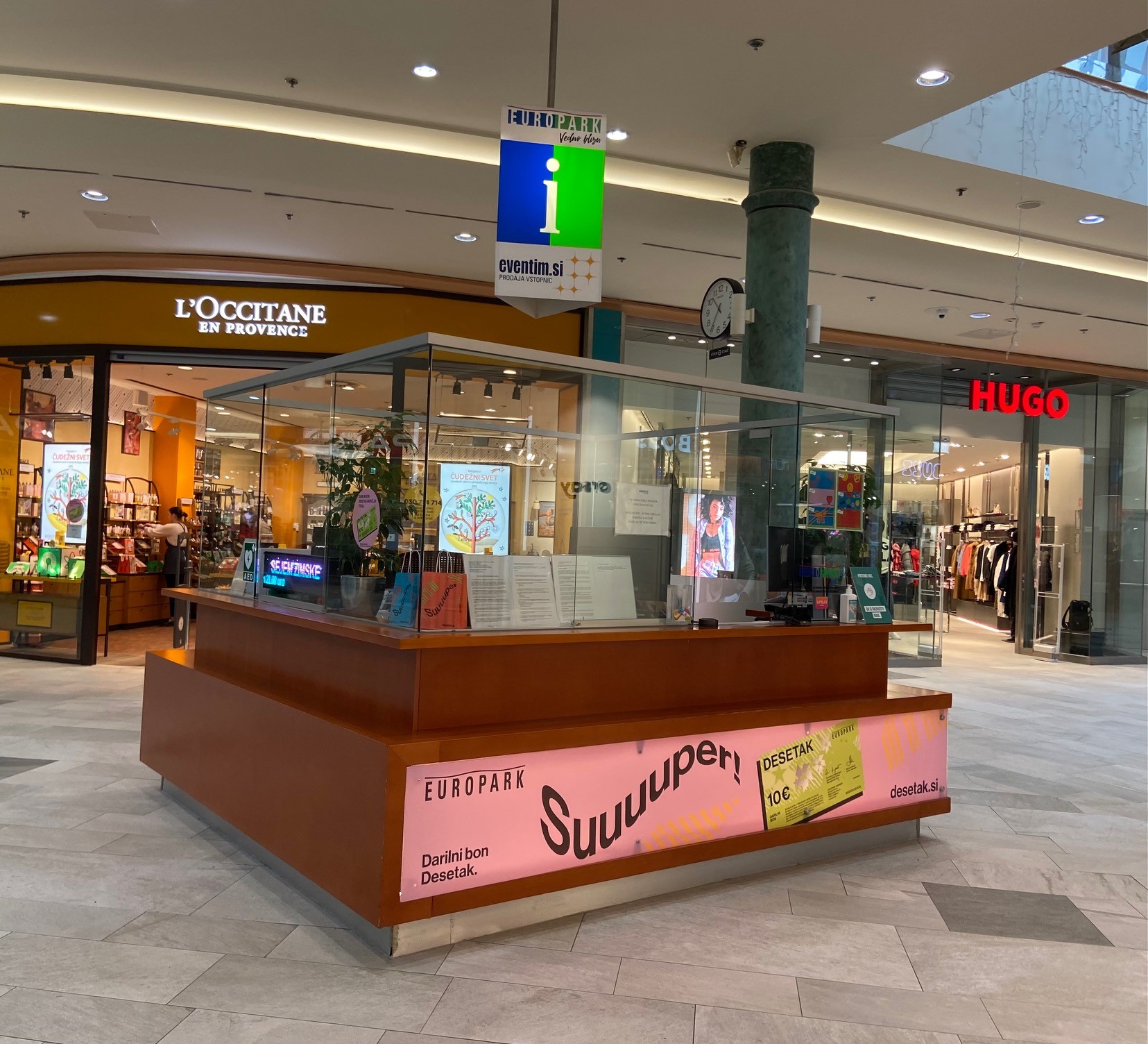 A shopping mall information desk with a colorful sign, surrounded by stores like L'Occitane and Hugo.