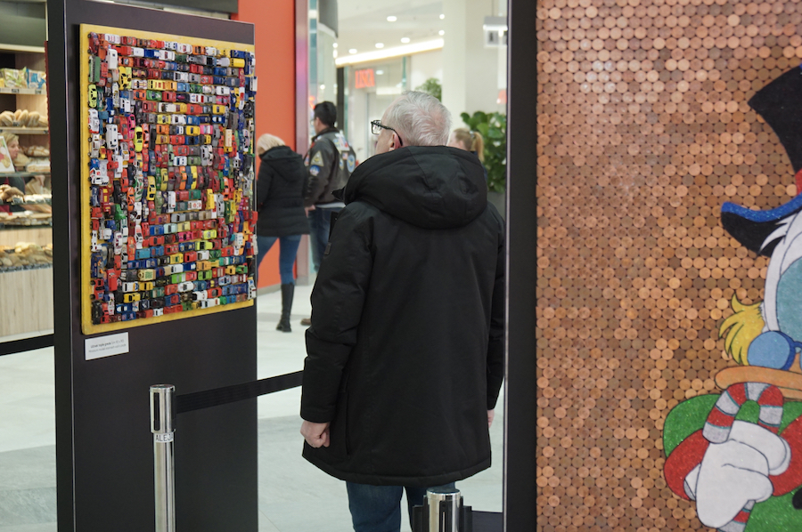 A man in a black coat observes a colorful art piece made of toy cars in a mall, with a cartoon character visible nearby.