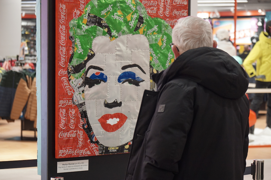 A man in a black coat admires a colorful artwork made from soda can labels, depicting a famous female figure.