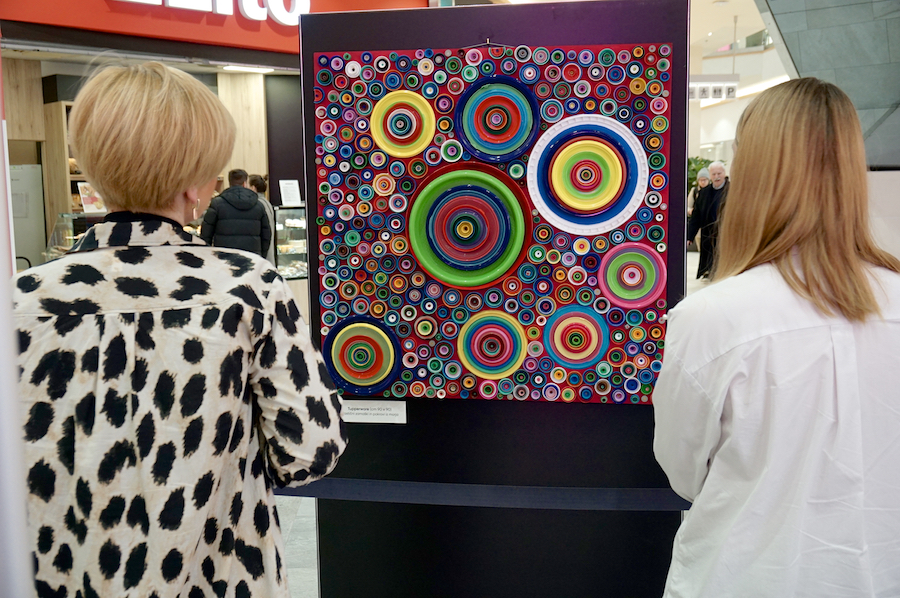 Two women observe a colorful circular artwork displayed in a gallery, surrounded by a modern interior.