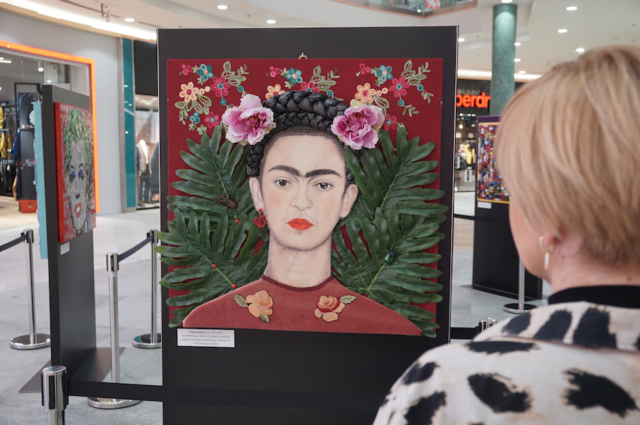 A person admires a vibrant artwork of a woman surrounded by flowers and leaves, displayed in a gallery setting.