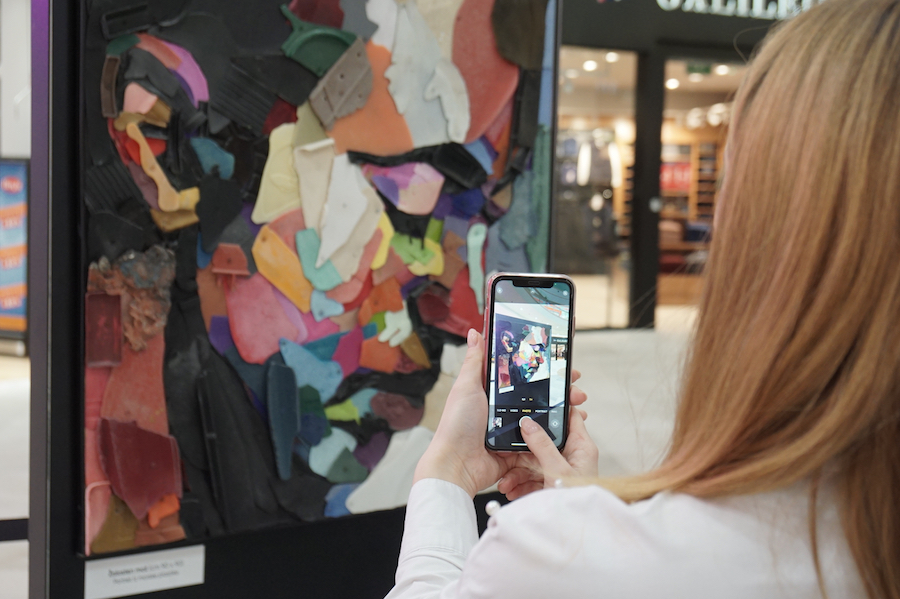 A person takes a photo of a colorful, abstract art piece made from various materials in a shopping area.