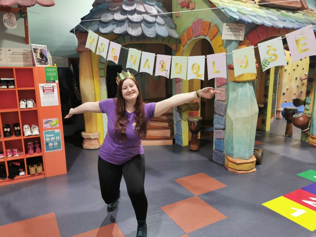 A smiling woman in a purple shirt poses playfully in a colorful indoor play area with whimsical structures and decorations.