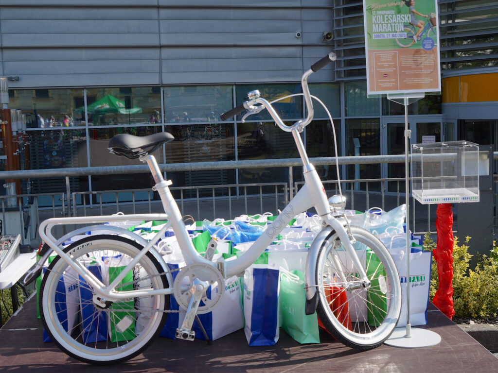 A white bicycle is displayed in front of colorful bags, with a sign for a cycling marathon in the background.