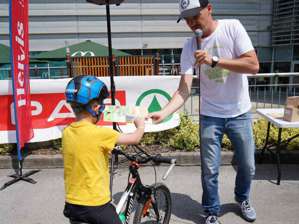 A child in a yellow shirt and helmet receives a certificate from a man with a microphone at an outdoor event.