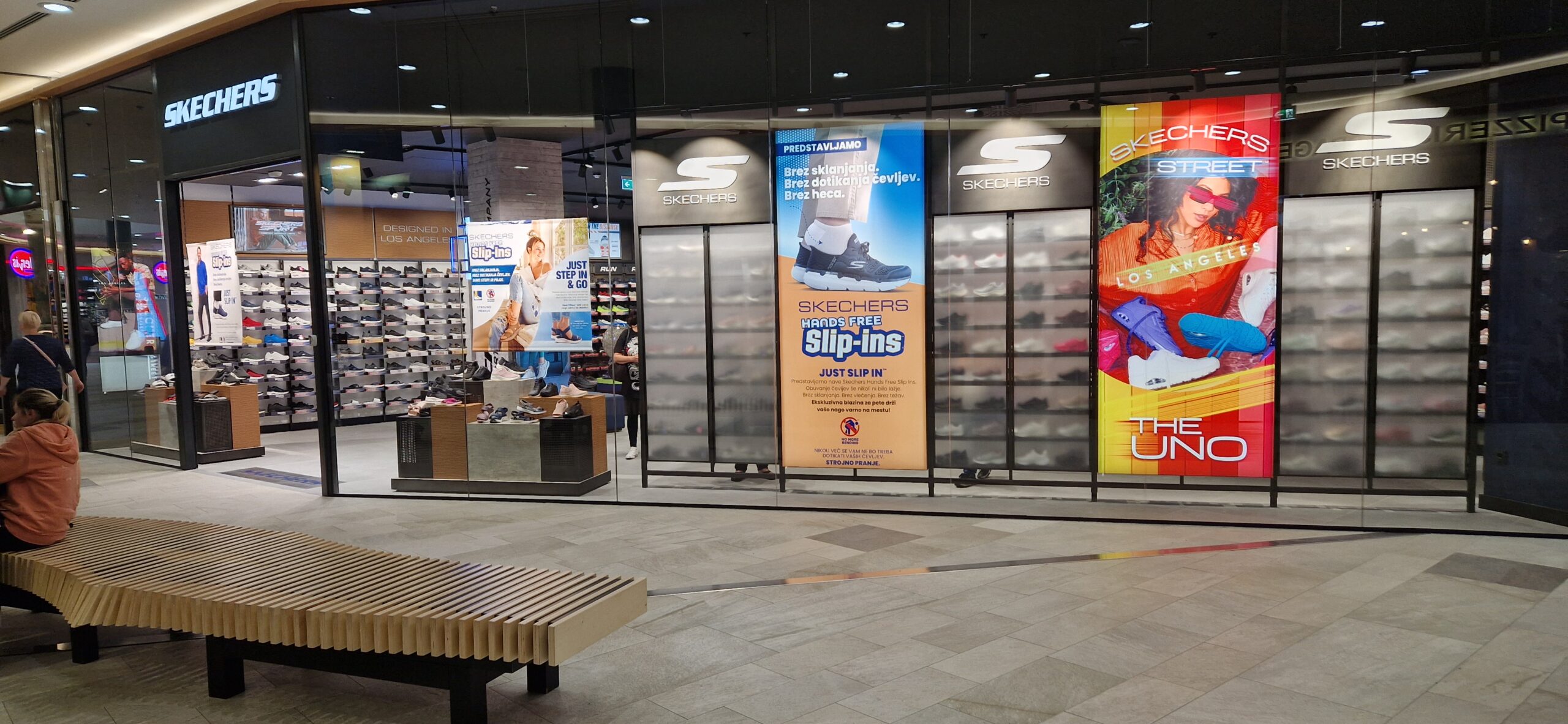 A Skechers store entrance featuring shoe displays and promotional banners for slip-ons and the UNO collection. A bench is visible in front.